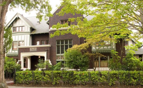 A large, historic brick house with green accents, surrounded by trees and a manicured garden.