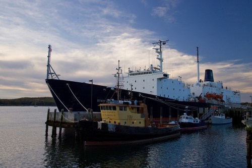 A large ship docked at a pier with a smaller tugboat nearby, under a cloudy sky.