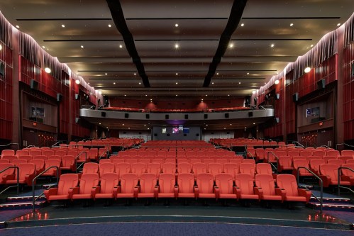 Interior of a modern theater with red seating, a spacious stage, and soft lighting, creating an inviting atmosphere.