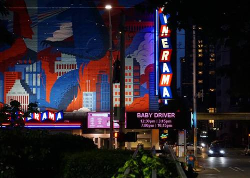 Neon-lit theater marquee displaying "Baby Driver" showtimes against a colorful mural backdrop at night.