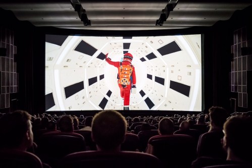 A cinema audience watches a scene from a space-themed film featuring an astronaut in a futuristic spacecraft.