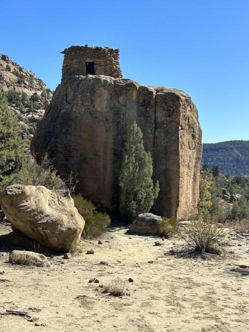 A rocky outcrop with a small stone structure on top, surrounded by sparse vegetation and a clear blue sky.
