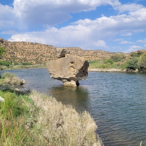 A large rock stands in the middle of a river, surrounded by greenery and hills under a blue sky with clouds.