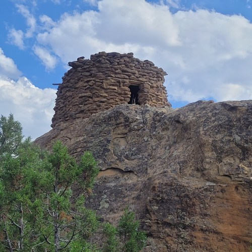A stone structure atop a rocky hill, surrounded by greenery and under a partly cloudy sky.