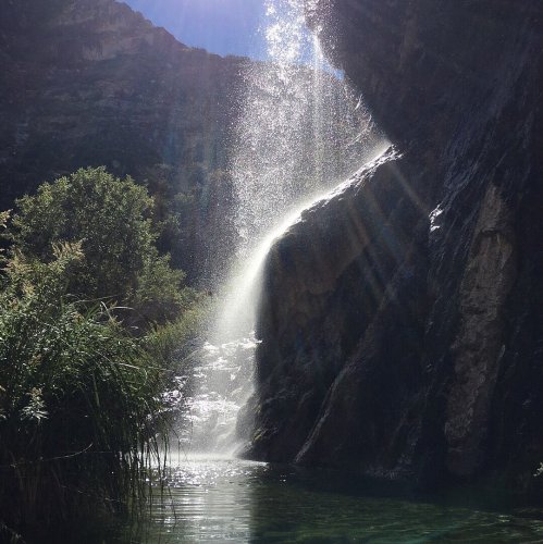 A waterfall cascades down rocky cliffs into a serene pool, surrounded by lush greenery and sunlight filtering through.