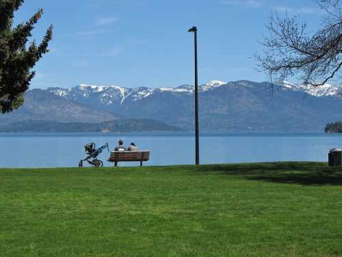 A couple sits on a bench by a lake, with mountains and a clear blue sky in the background. A stroller is nearby.