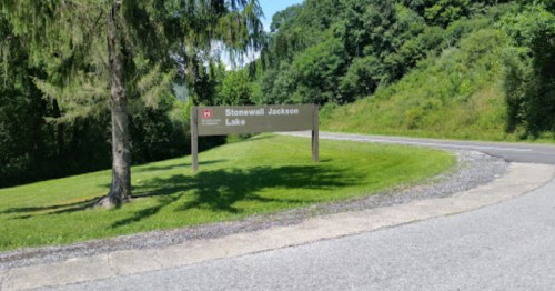 Sign for Stonewall Jackson Lake, surrounded by greenery and trees, near a winding road.