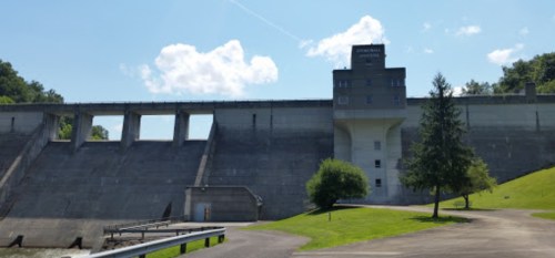 A concrete dam with a control tower, surrounded by green grass and trees under a blue sky with scattered clouds.