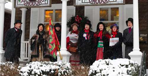 A group of people in winter attire singing carols on a snowy porch, surrounded by festive decorations.