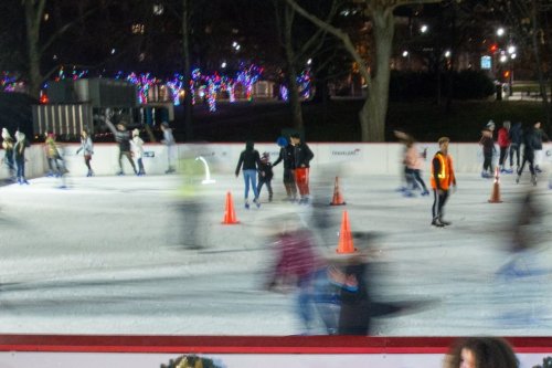 A busy ice skating rink at night, with skaters moving past orange cones and festive lights in the background.
