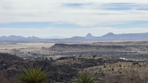 A panoramic view of a desert landscape with distant mountains and a clear sky, featuring rocky terrain and sparse vegetation.