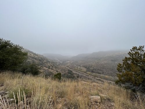 A misty valley view with rolling hills, sparse vegetation, and a winding road in the distance.