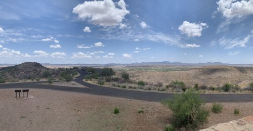 Panoramic view of a desert landscape with rolling hills, a winding road, and scattered clouds in a blue sky.
