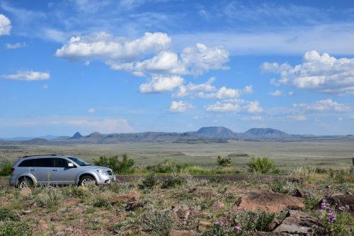 A silver SUV parked on a roadside with a vast desert landscape and mountains under a blue sky with fluffy clouds.