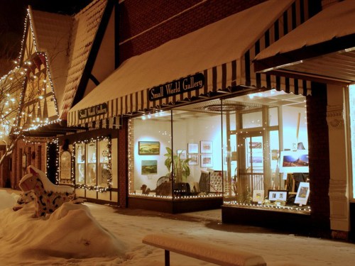 A cozy gallery with a striped awning, decorated with lights, showcasing art in its windows, surrounded by snow.