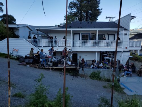 A band performs on an outdoor stage while an audience watches from a nearby area, surrounded by trees and buildings.