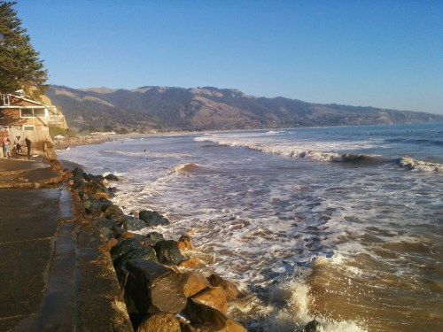 A scenic beach view with waves crashing on the shore, rocky coastline, and hills in the background under a clear blue sky.