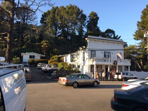 A quaint building with a porch, surrounded by trees and parked cars, with people gathered outside in the evening light.