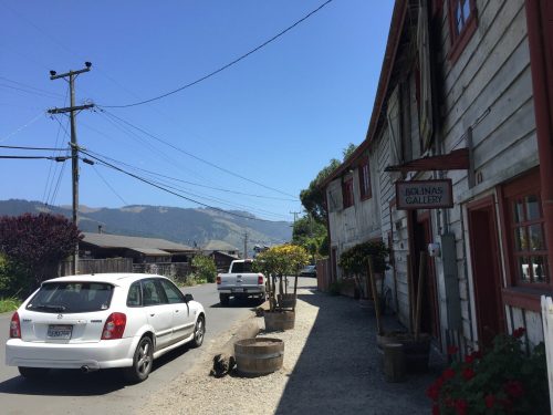 A sunny street scene in Bolinas, California, featuring parked cars and a gallery with mountains in the background.