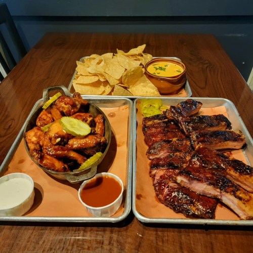 A platter featuring chicken wings, ribs, tortilla chips, and cheese dip on a wooden table.