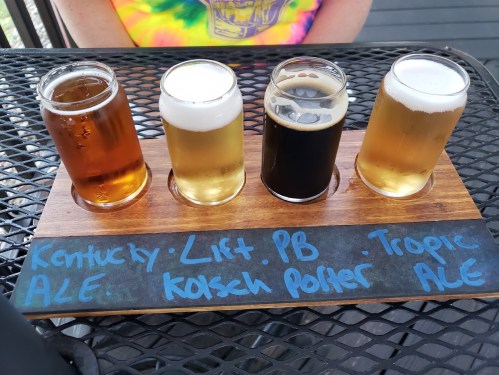A wooden flight board with four beer samples labeled Kentucky Ale, Lift PB, Kölsch Porter, and Tropic Ale.