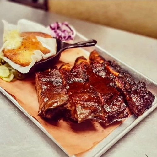 A platter featuring smoked ribs with barbecue sauce, cornbread, pickles, and coleslaw on a metal tray.