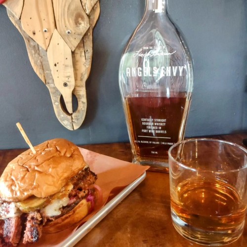 A close-up of a burger on a plate next to a glass of whiskey and a bottle of Angel's Envy bourbon.