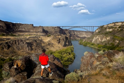 A person in a red jacket stands on a rocky ledge, overlooking a river and a bridge in a canyon landscape.