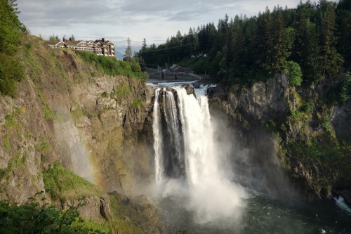 A majestic waterfall cascades down rocky cliffs, surrounded by lush greenery and a distant building.