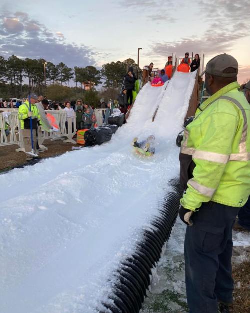A snowy slide with people sledding down, surrounded by spectators and a worker in a safety vest.