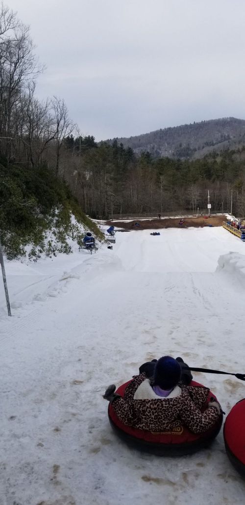 A snowy hill with people tubing down, surrounded by trees and mountains in the background.