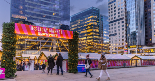People walk past the entrance of a festive holiday market, surrounded by twinkling lights and modern buildings.