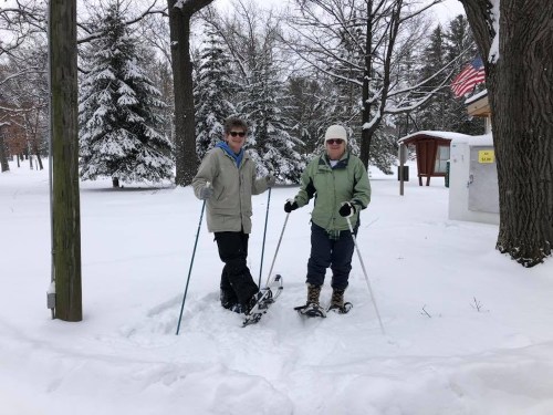 Two people stand in a snowy landscape, wearing winter gear and snowshoes, surrounded by trees and a snowy path.