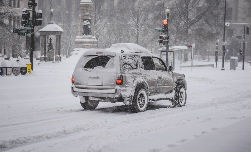 A vehicle driving through a snowy street, with heavy snowfall and traffic lights visible in the background.