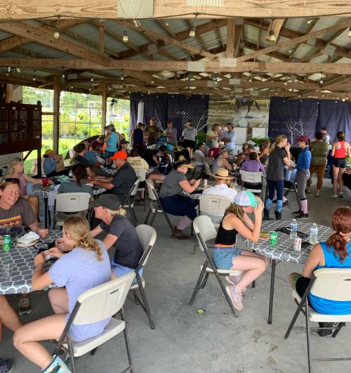 A large group of people dining and socializing in a covered outdoor space, with tables and chairs arranged throughout.