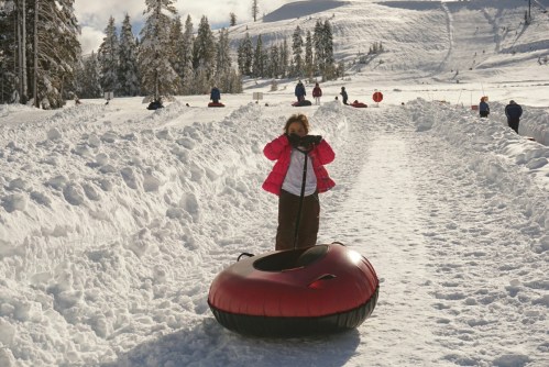 A child in a pink jacket stands next to a red snow tube on a snowy slope, with other people tubing in the background.