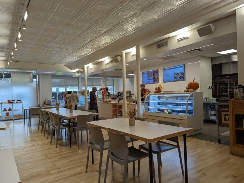 Bright, modern café interior with tables, a display case, and customers at the counter. Warm, inviting atmosphere.