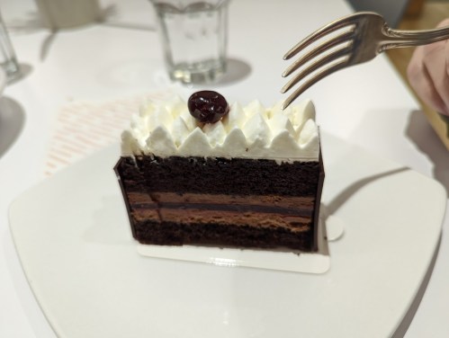 A slice of layered chocolate cake topped with whipped cream and a cherry, with a fork poised to take a bite.