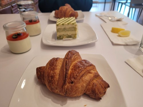 A croissant in the foreground, with a green cake and desserts in jars on white plates in the background.