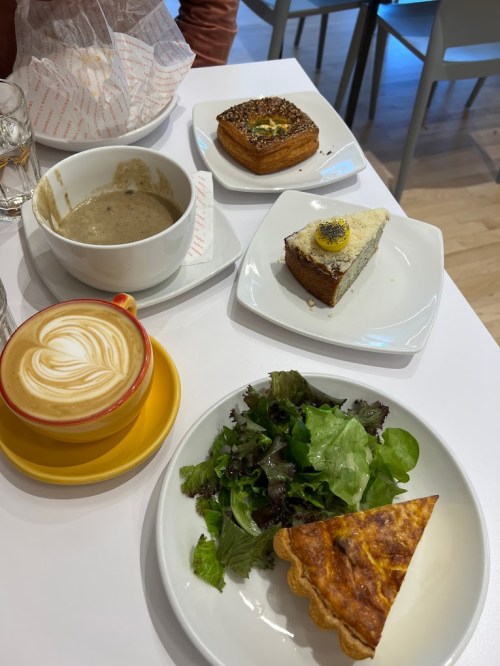 A table with a latte, salad, quiche, soup, and pastries on white plates.