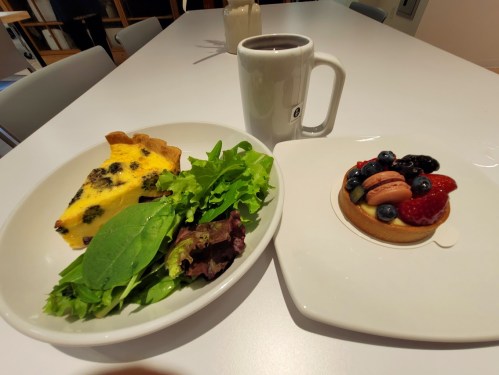 A plate with a slice of quiche, mixed greens, and a dessert tart topped with berries and macarons, alongside a coffee mug.