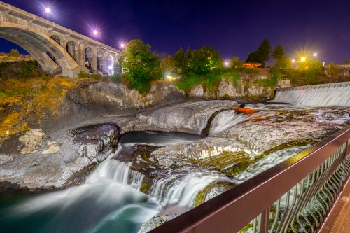 A serene night scene of a waterfall under a bridge, illuminated by lights, with flowing water and rocky surroundings.