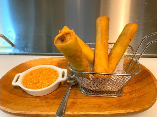 Crispy spring rolls in a metal basket with a side of dipping sauce on a wooden plate.