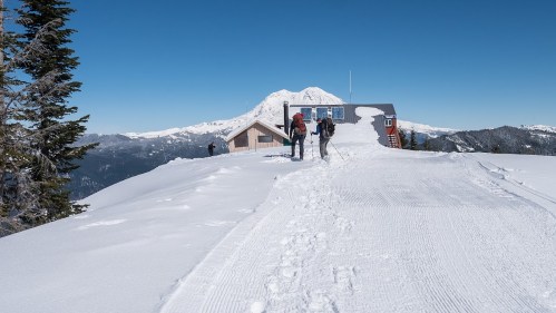 Two hikers trek through snow towards a mountain lodge, with a snow-covered peak in the background under a clear blue sky.