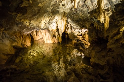 A dimly lit cave with stalactites and stalagmites, featuring a calm pool of water reflecting the rocky surroundings.