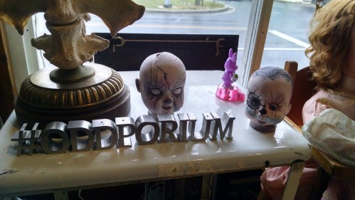 Two creepy doll heads on a table with a sign that reads "#ODDPORIUM" in front of them.