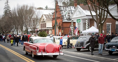 A festive street scene with vintage cars, holiday decorations, and crowds enjoying a winter event. Snow on rooftops.