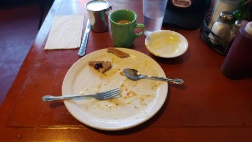 A nearly empty plate with remnants of food, a spoon, and a green cup on a wooden table.