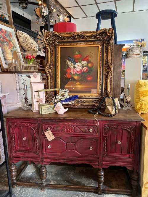 A vintage red cabinet displays a floral painting, decorative items, and a telephone in a cozy shop setting.