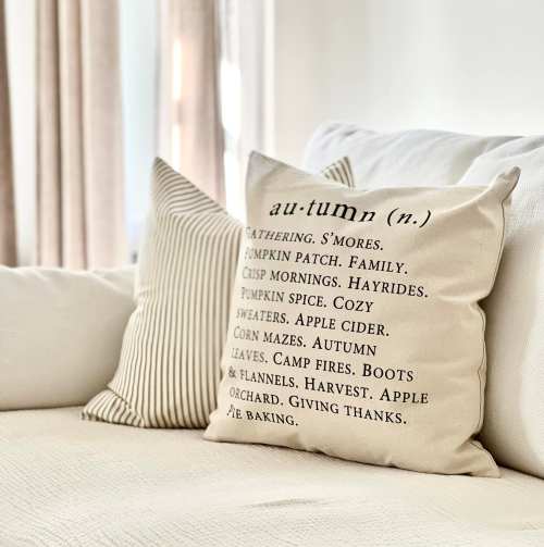A decorative pillow on a couch featuring autumn-themed words and phrases like "s'mores," "pumpkin patch," and "harvest."